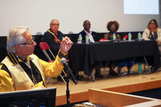 An Indigenous elder speaks into a microphone, gesturing with his hand during a panel discussion. Four other panelists, including two women and two men of diverse backgrounds, sit at a table in the background.