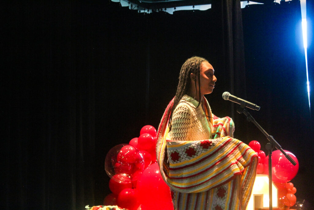 A Black woman stands at a microphone on stage, wrapped in a colourful blanket. She is delivering a speech or performance. In the background, bright red balloons and lights are visible, contrasting with the dark setting. Une femme noire se tient devant un micro sur scène, enveloppée dans une couverture multicolore. Elle prononce un discours ou fait une performance. En arrière-plan, des ballons rouges vifs et des lumières sont visibles, contrastant avec le décor sombre.