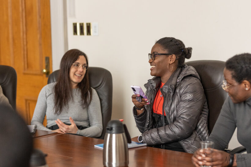 A group of people sitting around a conference table, engaging in discussion. One person is smiling while holding a phone, and another is listening attentively. - Un groupe de personnes assis autour d'une table de conférence, participant à une discussion. Une personne sourit en tenant un téléphone, tandis qu'une autre écoute attentivement.